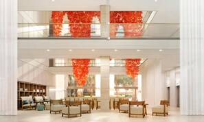 A spacious lobby with a high ceiling, a seating area with wooden chairs and tables, and a bookshelf filled with books. - Hilton Barcelona (Barcelona)