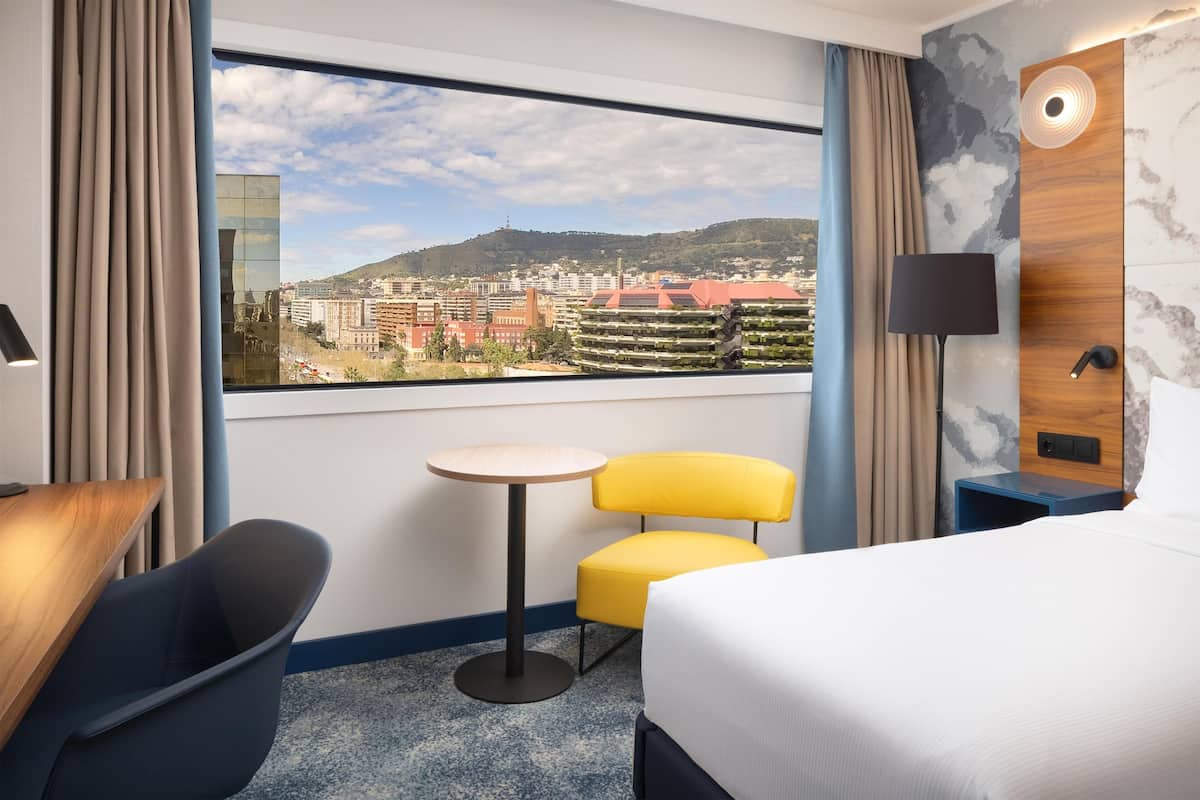 A hotel room with a bed, a small round table, a yellow chair, a blue chair, and a view of a cityscape through a large window.