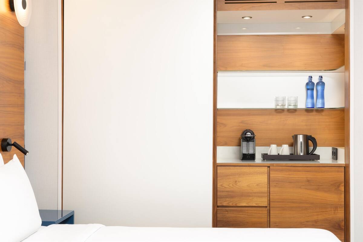 A hotel room with a bed, a nightstand, a wooden wardrobe, a shelf with bottles and glasses, and a coffee maker.