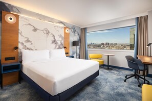 A hotel room with a large bed, a desk, a chair, a small table, and a view of the city. - Hilton Barcelona (Barcelona)