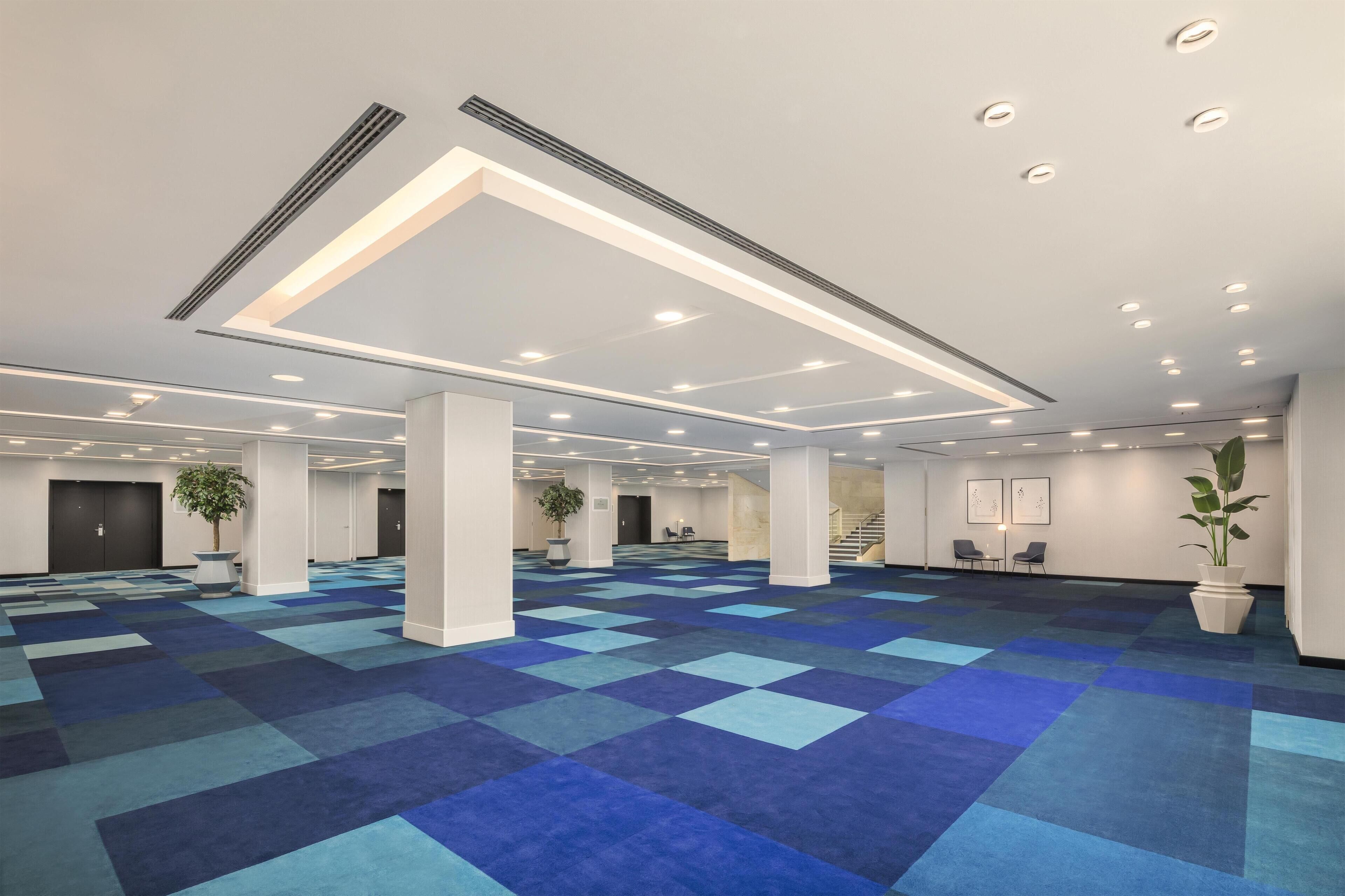 A spacious hotel lobby with a checkered blue carpet, recessed lighting, and potted plants.