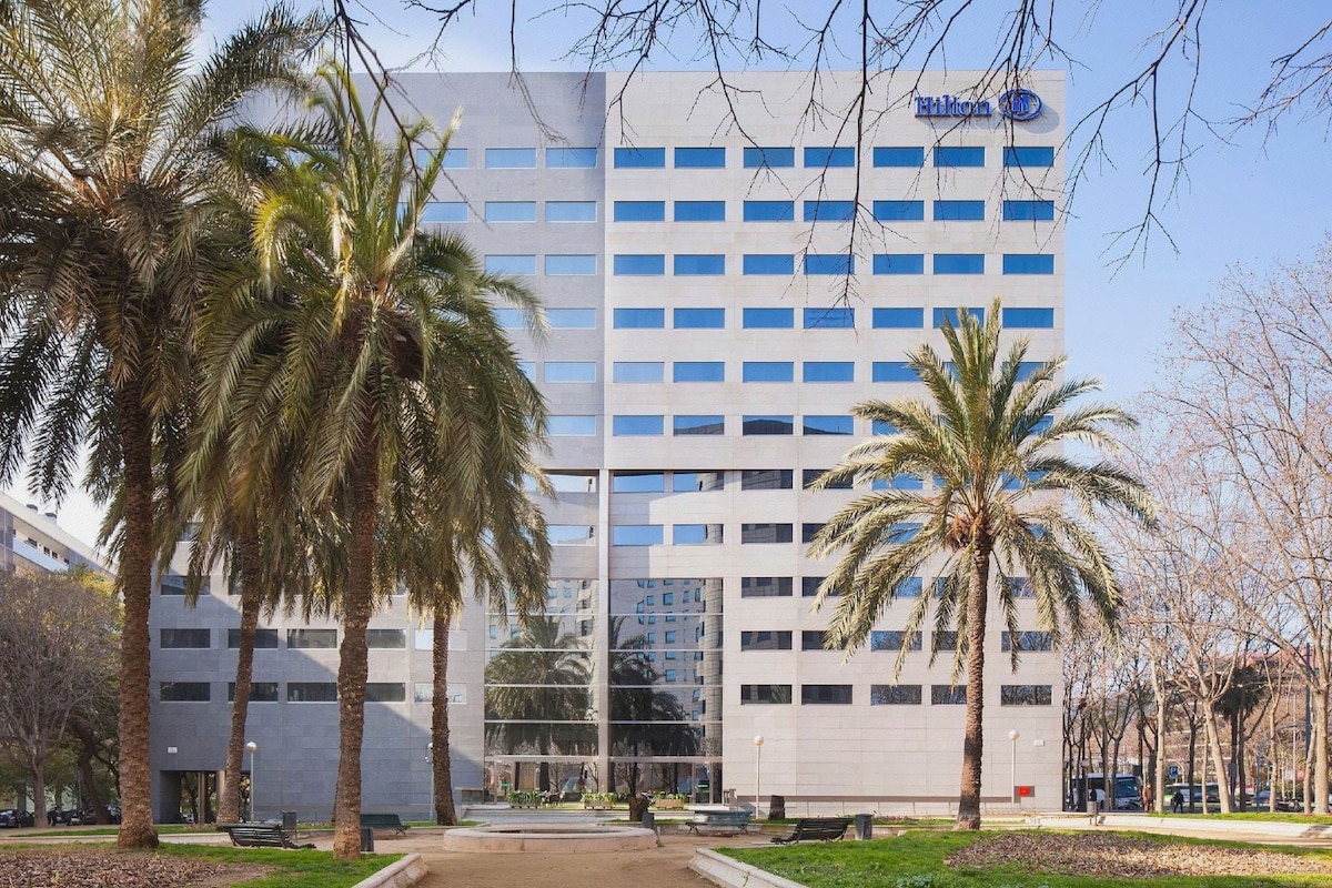 A modern building with a glass facade, palm trees, and a clear sky.