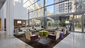 Un hall d’entrée spacieux au design moderne, comprenant différents espaces de détente, un coin café et de grandes fenêtres offrant une vue sur le paysage urbain.