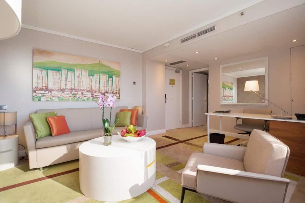 A modern hotel room with a sofa, a round coffee table with fruit, a painting on the wall, and a desk with a lamp.