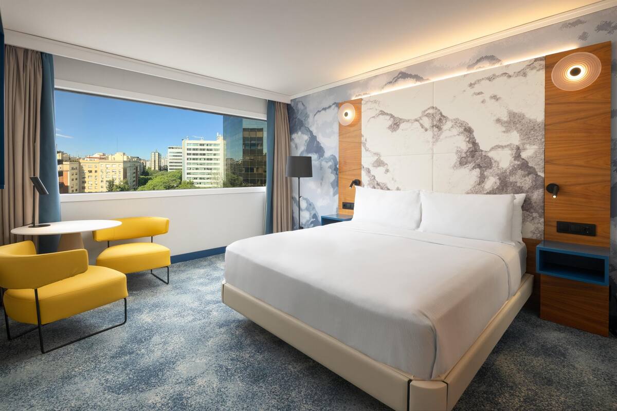 A hotel room with a large bed, a small table with yellow chairs, a city view, and a marble-patterned wall.