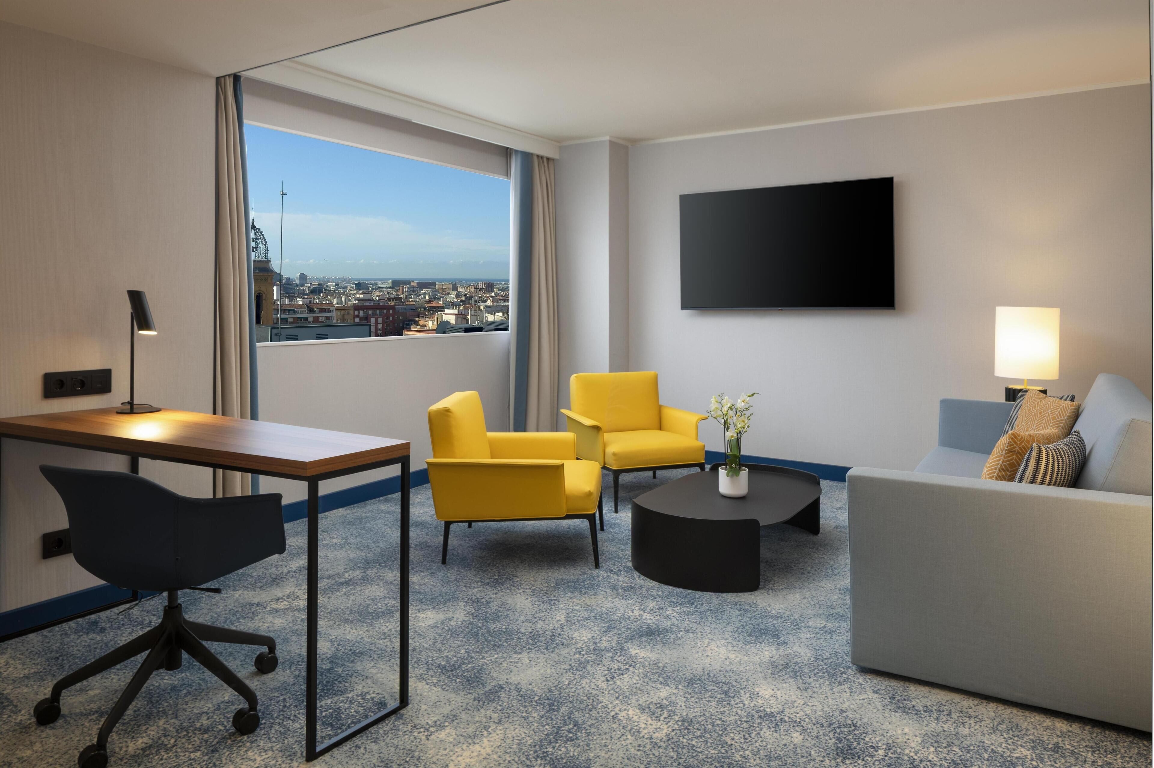 Une chambre d’hôtel équipée d’un bureau, de deux fauteuils jaunes, d’un canapé gris, d’une table basse noire et d’un grand téléviseur fixé au mur.