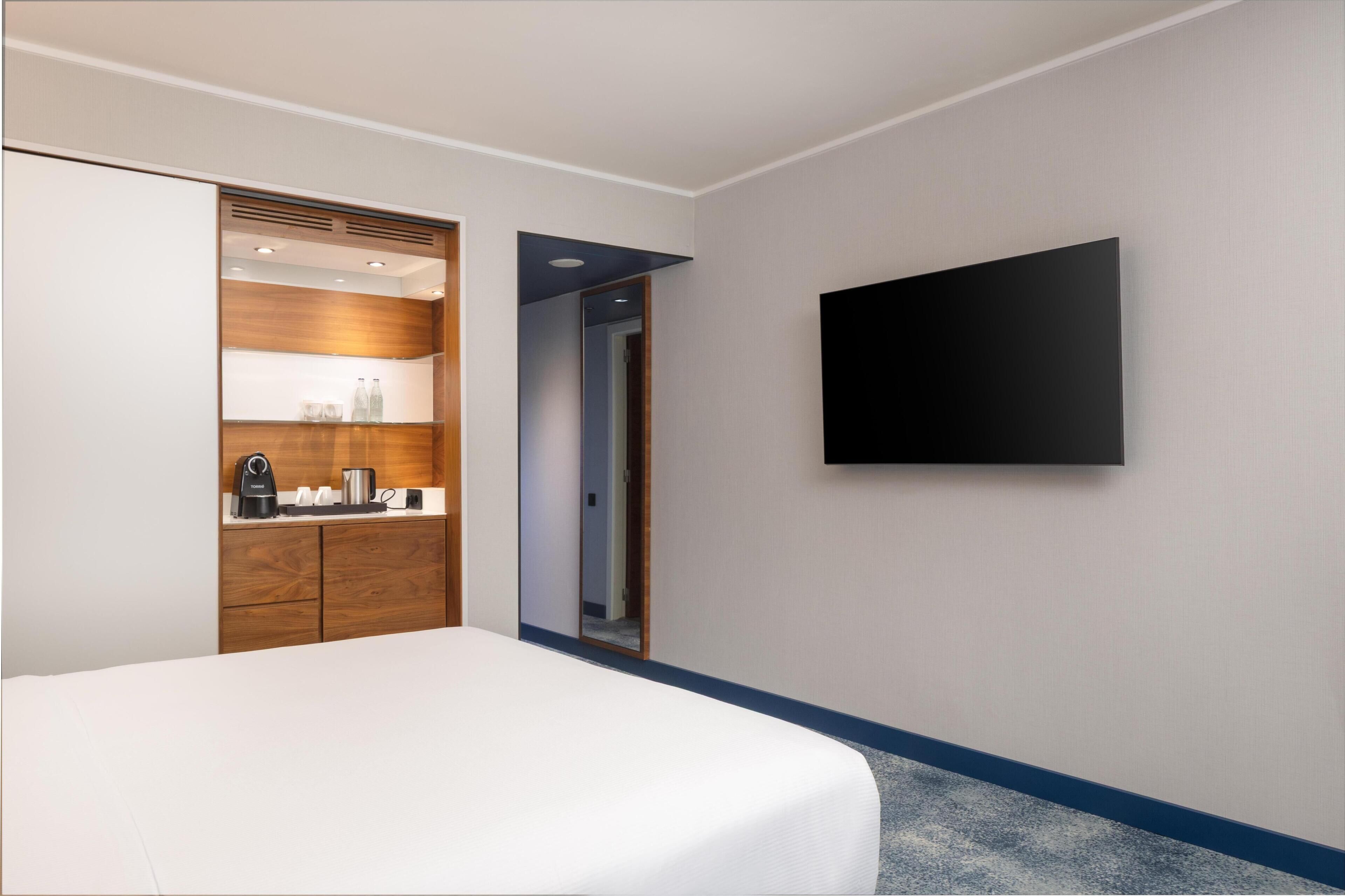 A hotel room with a bed, a wardrobe with a mirror, a flat-screen TV mounted on the wall, and a small kitchenette area.