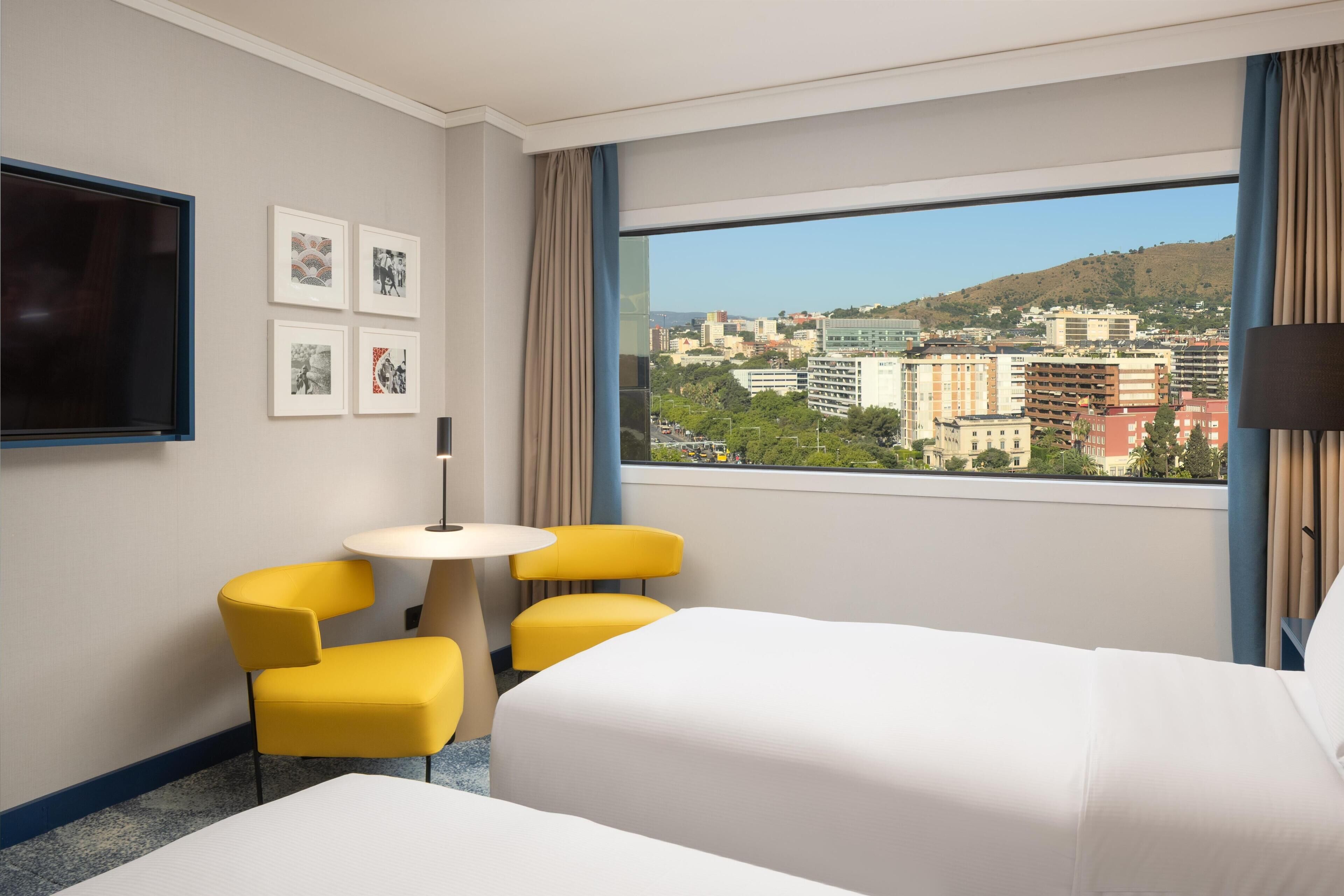 A hotel room with a bed, a TV, a small table with two chairs, and a city view through the window.