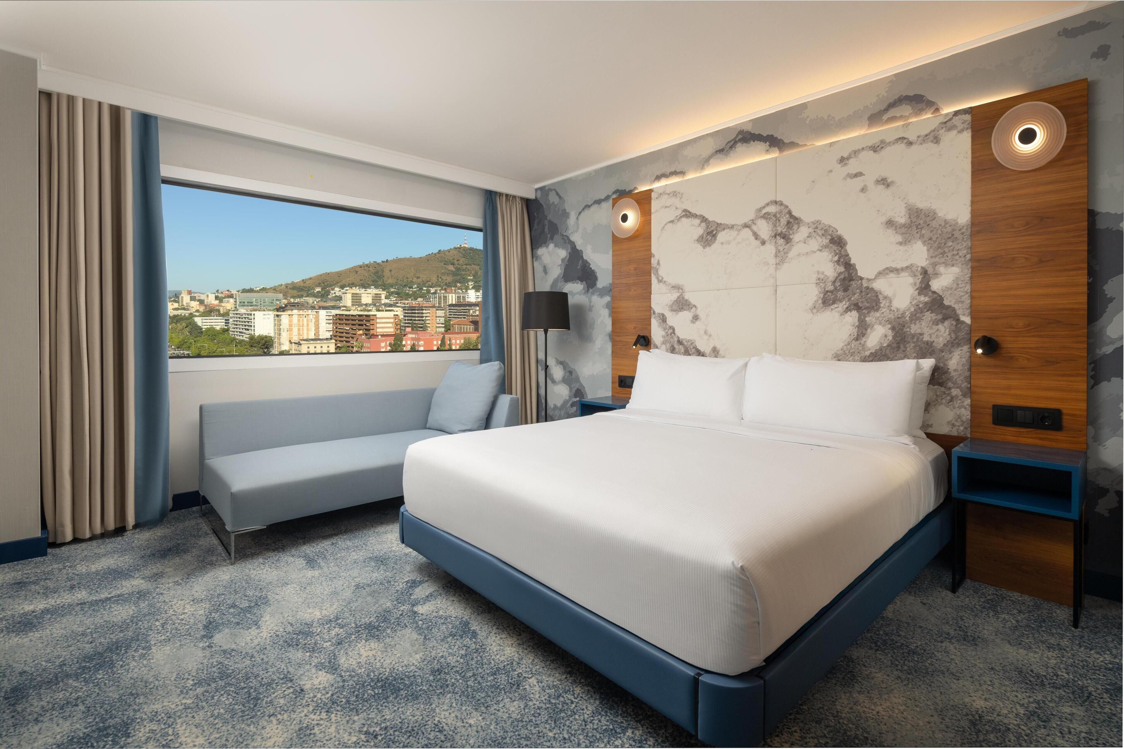 A hotel room with a bed, a sofa, a nightstand, a lamp, and a view of a cityscape.