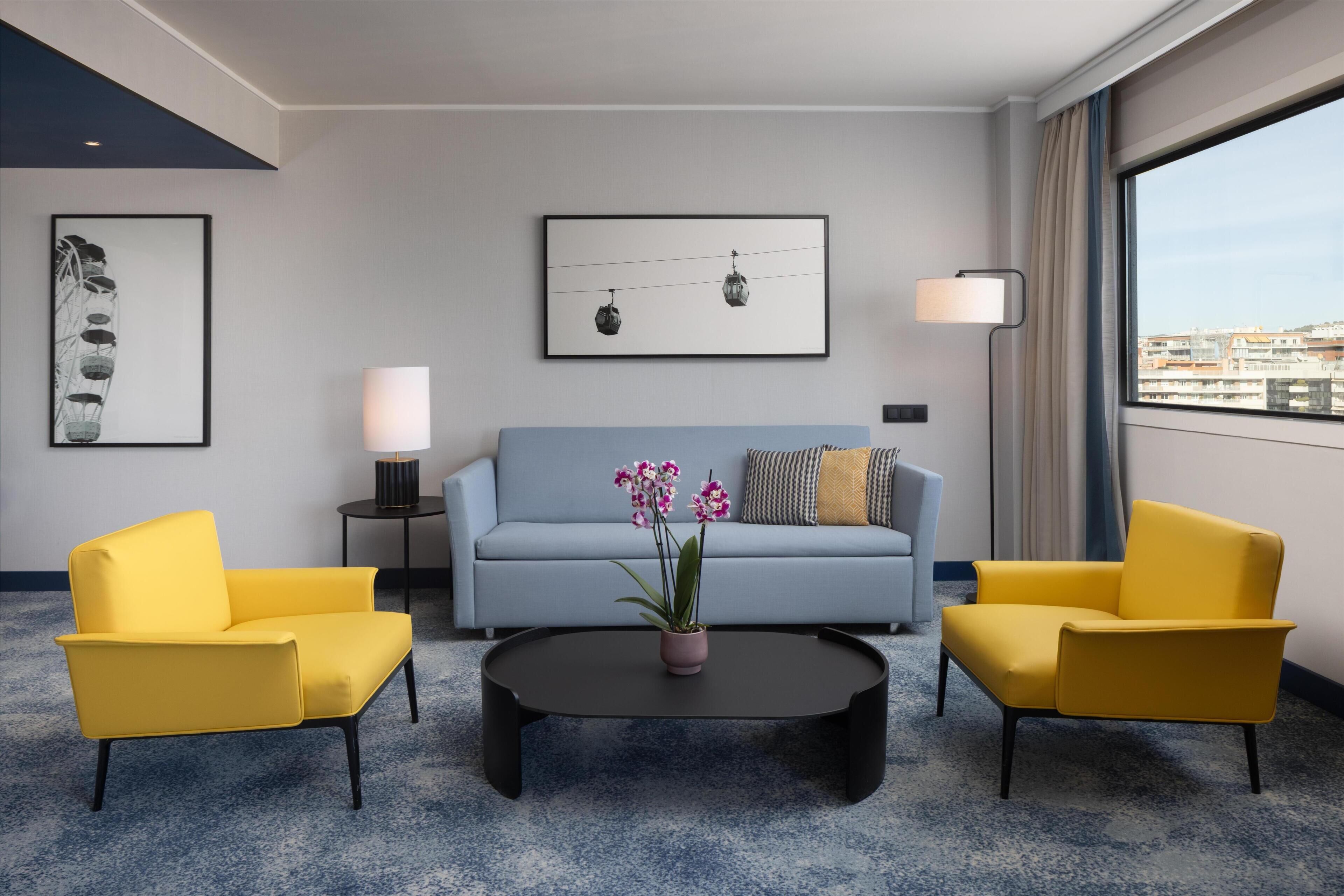 Hotelkamer met een blauwe bank, twee gele fauteuils, een bijzettafeltje en uitzicht op de stad.