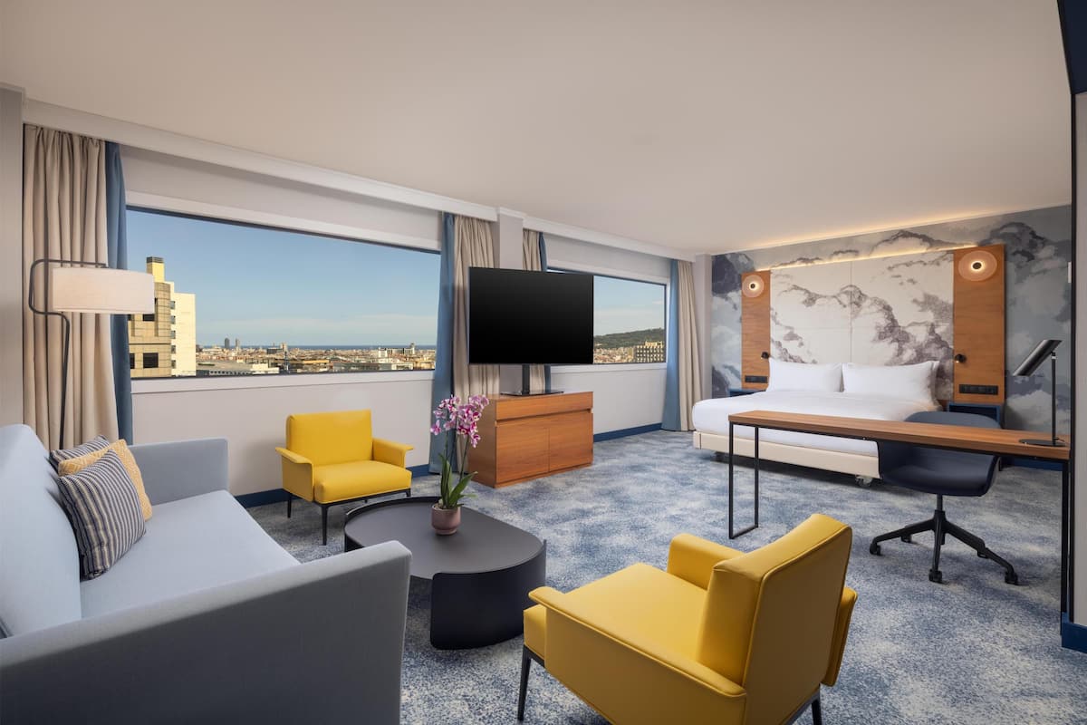 A hotel room with a city view, a bed, a desk, a TV, and a coffee table.