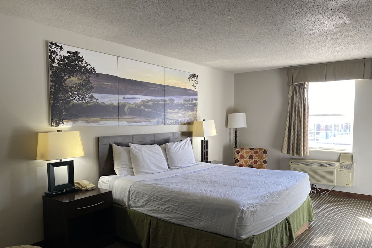 Een hotelkamer met een groot bed, twee bedlampen, een flatscreen-televisie, een bureau met telefoon, een stoel, een raam met gordijnen en een luchtverkoeler aan de muur.