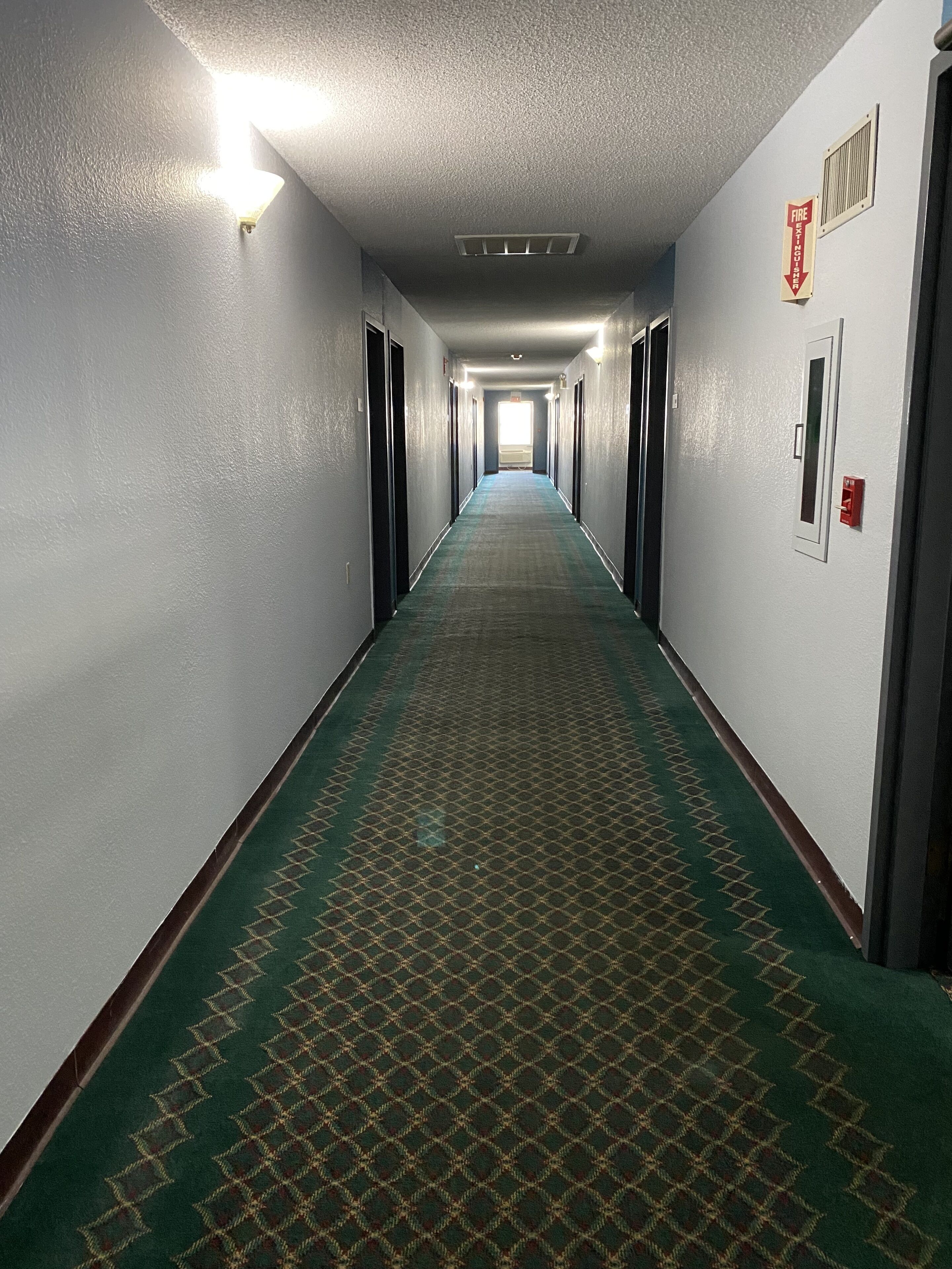 A long, narrow hallway with a green patterned carpet, white walls, and doors on both sides.