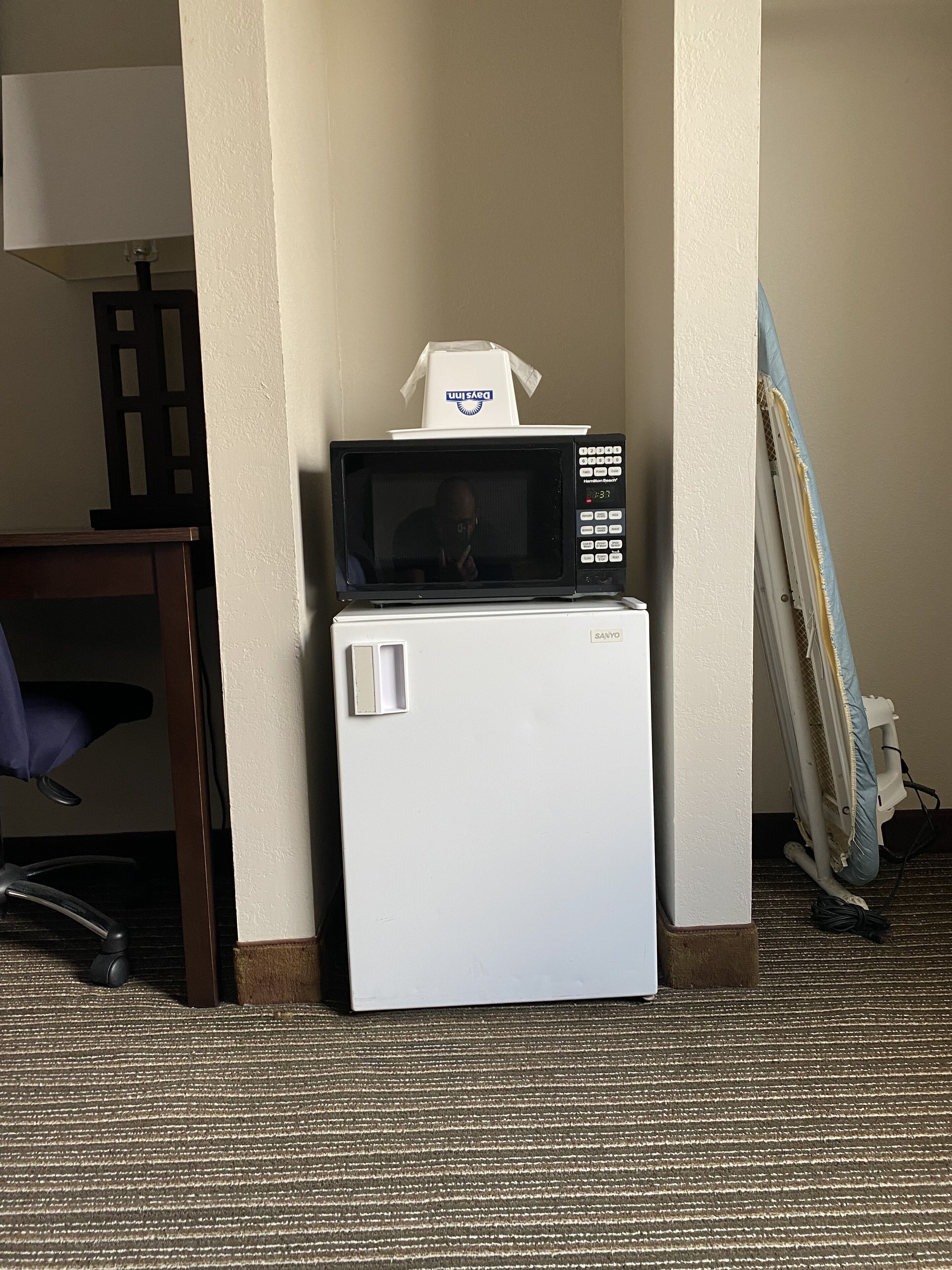 A compact kitchenette with a microwave and refrigerator, a chair, and a partially visible ironing board.