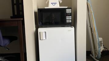 A compact kitchenette with a microwave and refrigerator, a chair, and a partially visible ironing board.