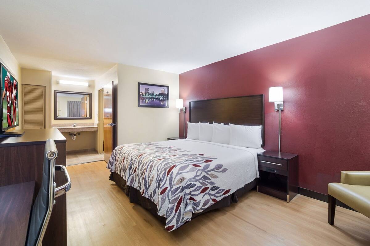 A hotel room with a bed, bedside table, lamp, chair, and a television on a desk.