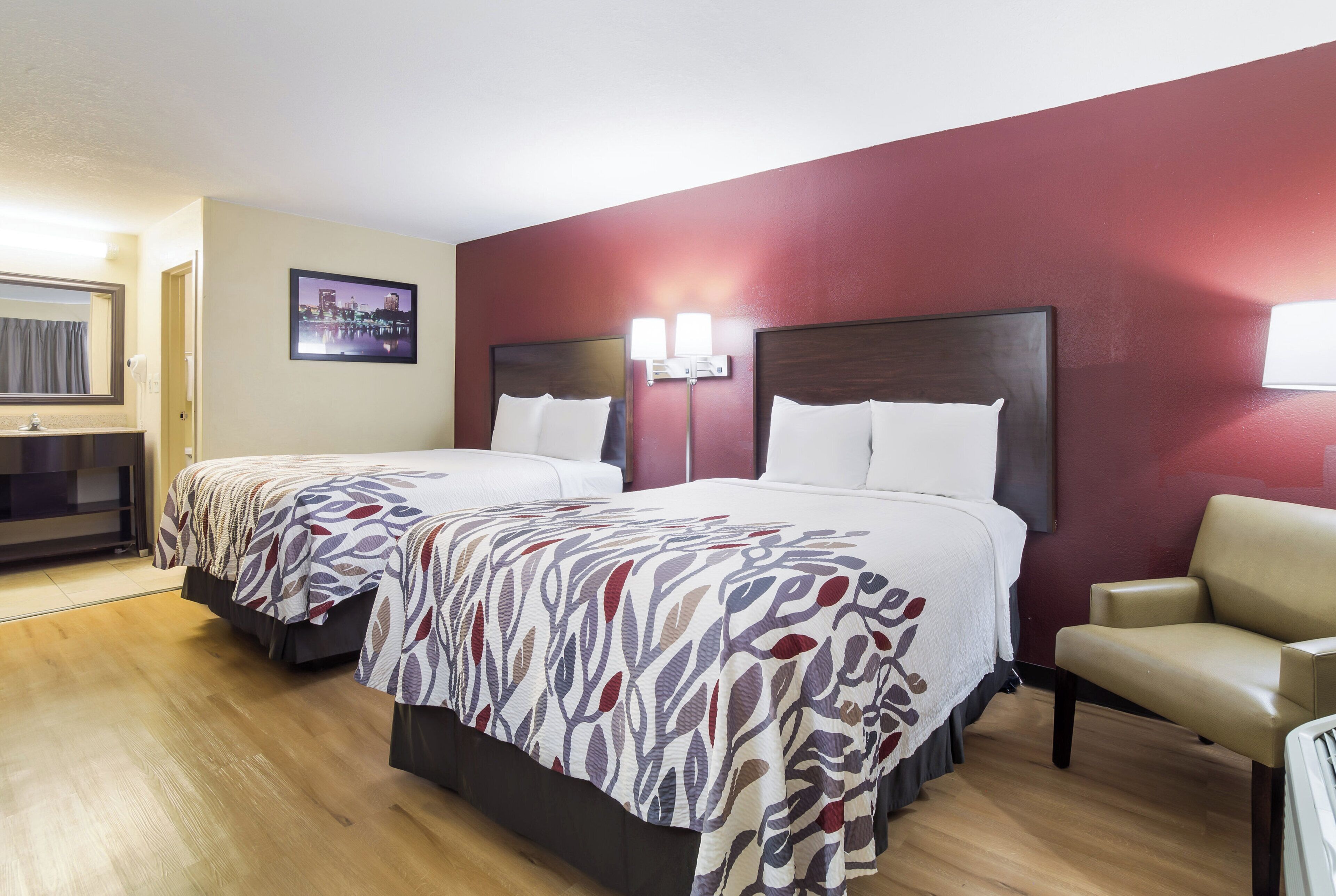 Une chambre d’hôtel avec deux lits, une chaise, un bureau et un tableau représentant un paysage urbain.