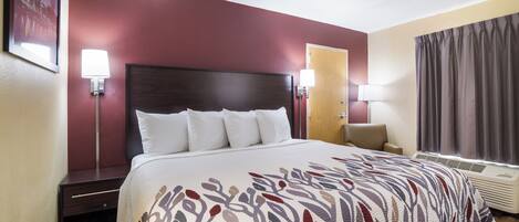 A hotel room with a large bed, a nightstand, a desk, a chair, a window with curtains, and wall-mounted lamps.