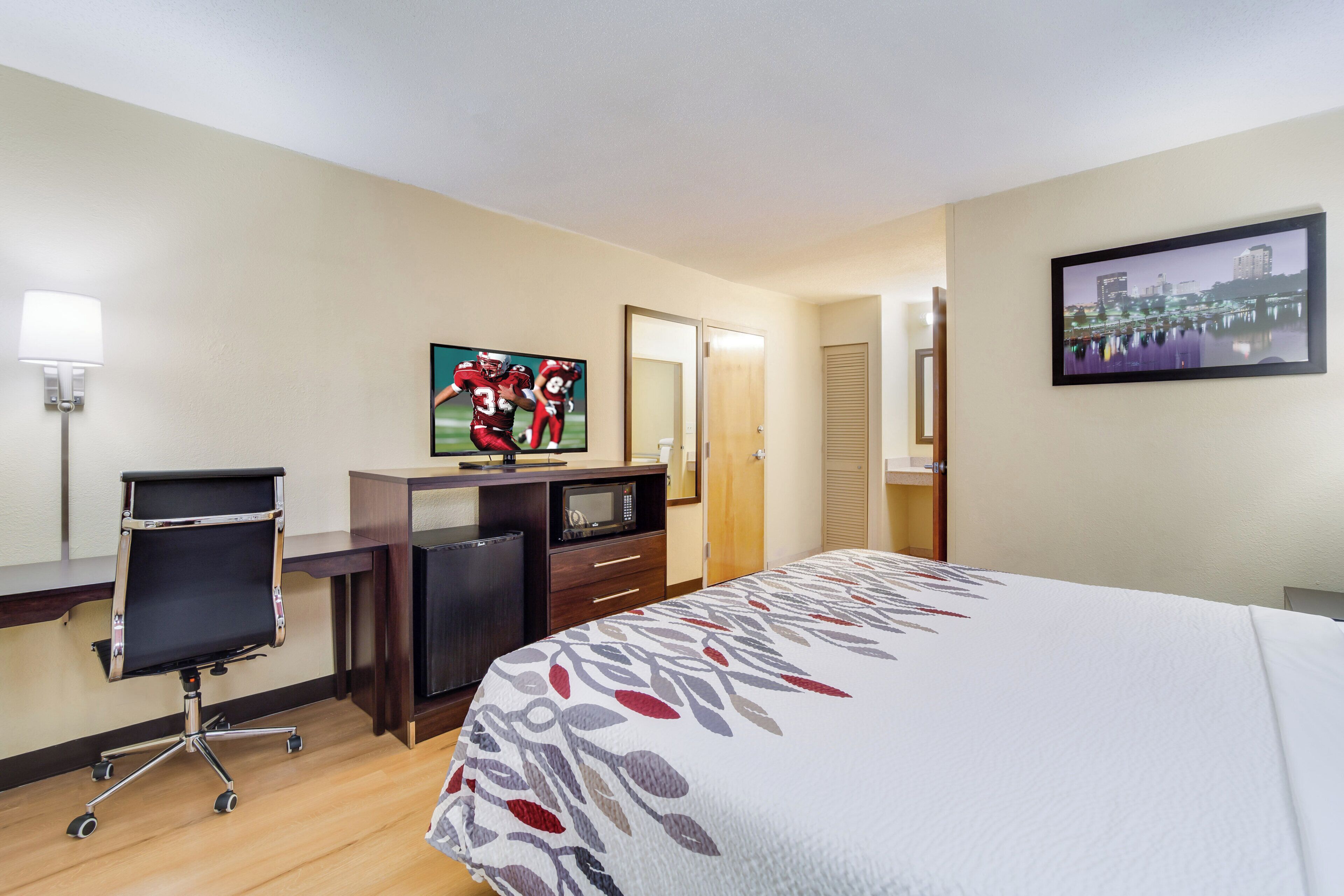 A hotel room with a bed, a desk with a computer, a TV, a mirror, and a cityscape picture on the wall.