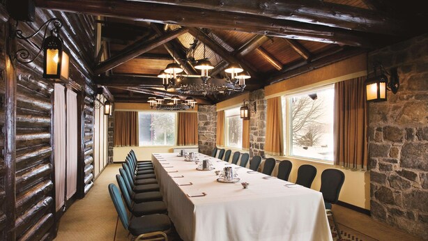 A long conference table with chairs, set against a stone wall and wooden beams, with large windows and hanging lights.