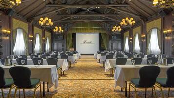 A banquet hall with tables set for a formal event, featuring chandeliers and a stage with a backdrop.