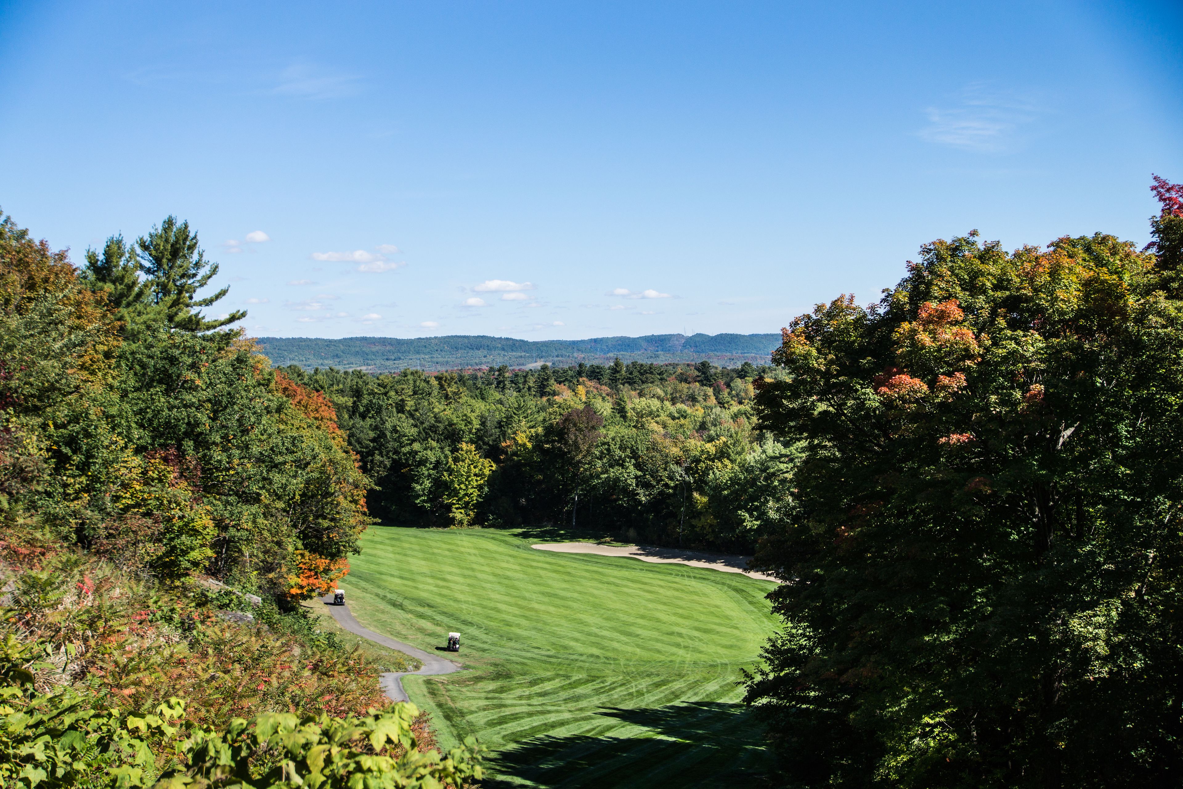 A golf course with a clear path, surrounded by dense trees and a distant view of hills.