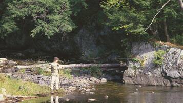 A person standing by a calm river with a rocky bank and dense trees in the background.