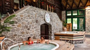 Zona de piscina interior con paredes de piedra, un reloj, cubierta de madera y un jacuzzi.