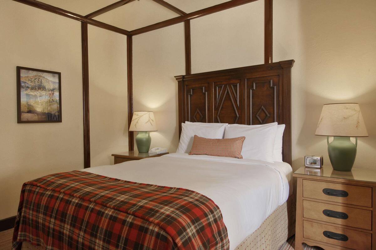 Een net opgemaakt bed met een geruit plaid, twee bedlampen en een nachtkastje met een telefoon en een klein horloge.
