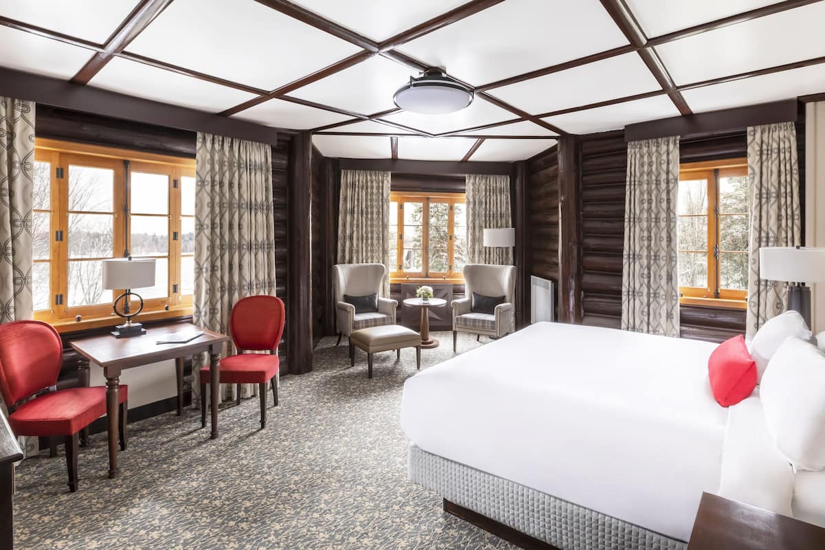 Een hotelkamer met een groot bed, twee fauteuils, een bureau en een kleine tafel. Er zijn twee ramen met gordijnen, een plafondlamp en uitzicht op bomen.