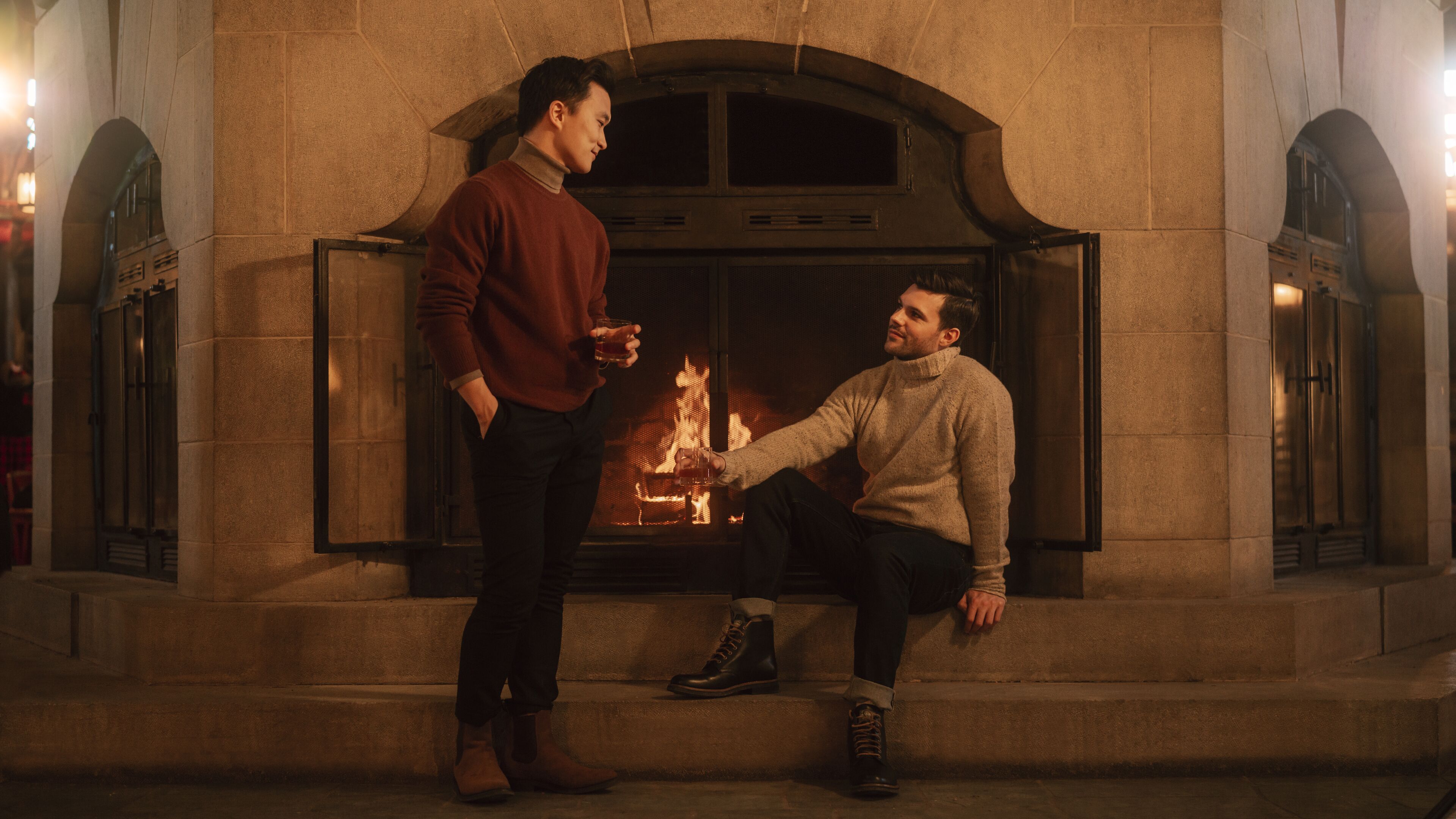 Dos personas sentadas junto a una chimenea en una habitación con paredes de piedra.