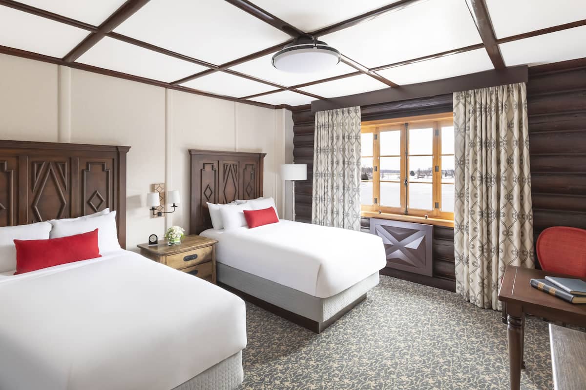 Een hotelkamer met twee bedden, een houten hoofdbord, een bureau met stoel, een lamp en een raam met gordijnen.