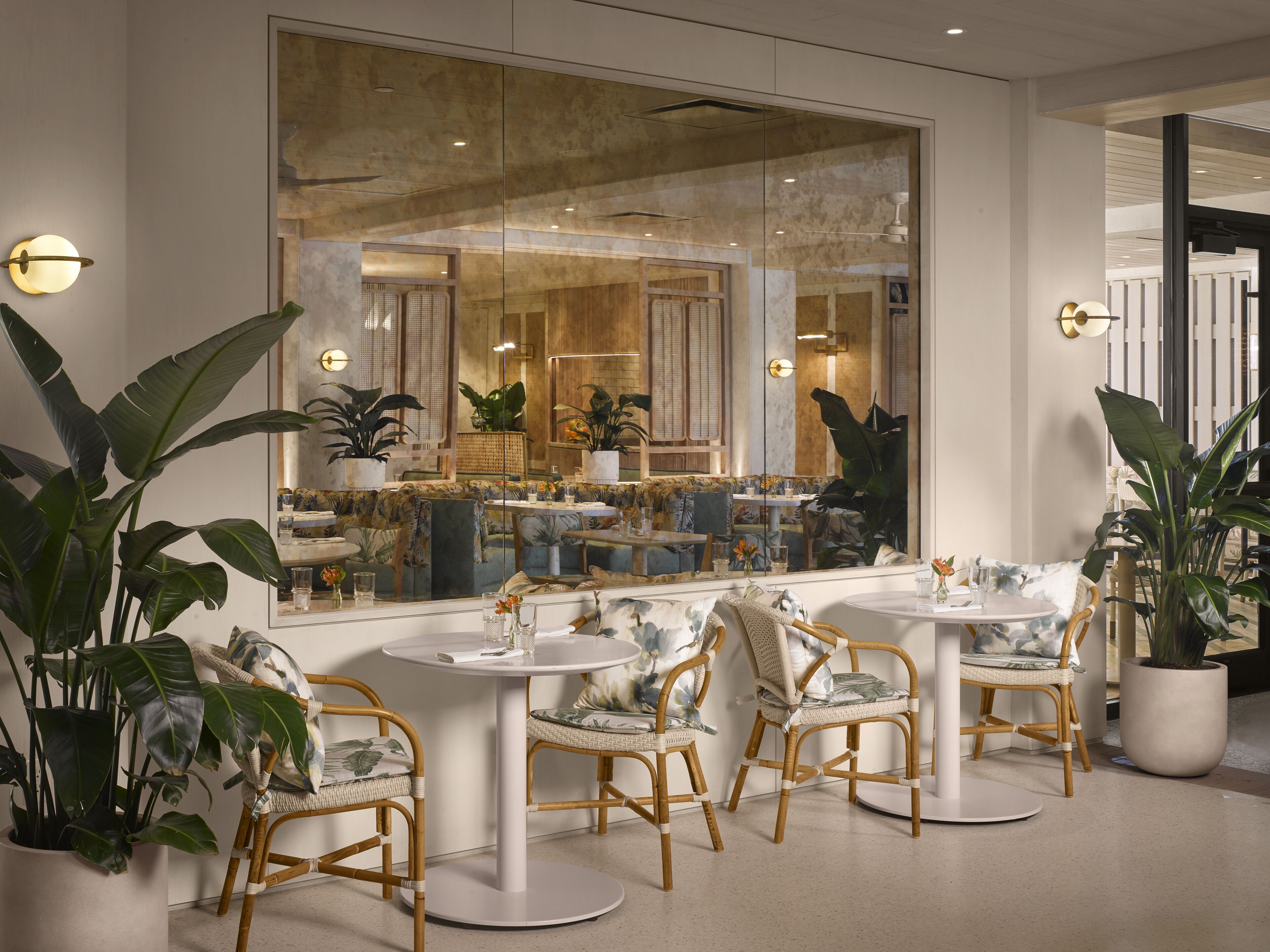 A modern interior with round tables and wicker chairs, large potted plants, and a large mirror reflecting the room.