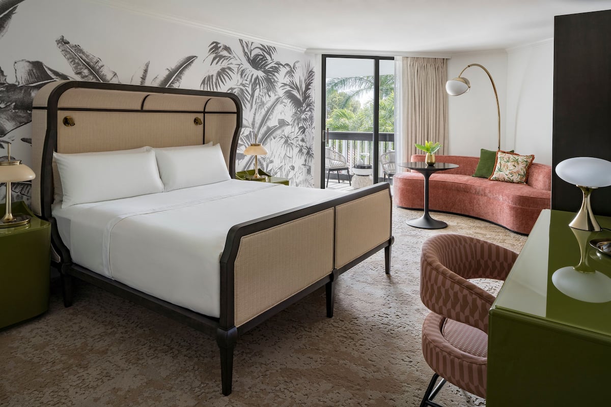 Hotelkamer met een bed, een bank, een stoel, een tafel en een balkon met uitzicht op palmbomen.