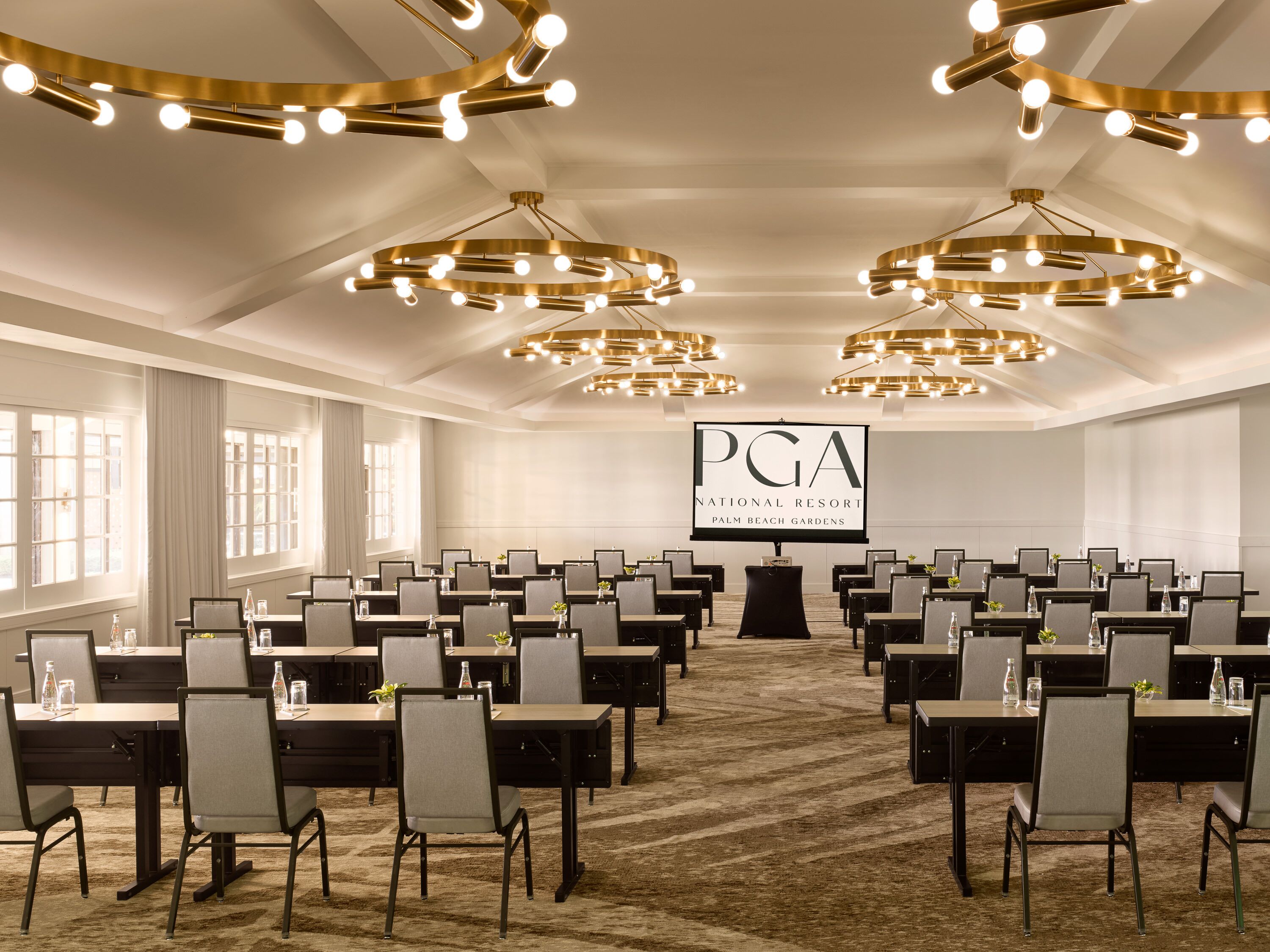 Ein Konferenzraum mit Tischen und Stühlen, die in Reihen angeordnet sind, einem großen Bildschirm, auf dem "PGA National Resort Palm Beach Gardens" angezeigt wird, und modernen Kronleuchtern.