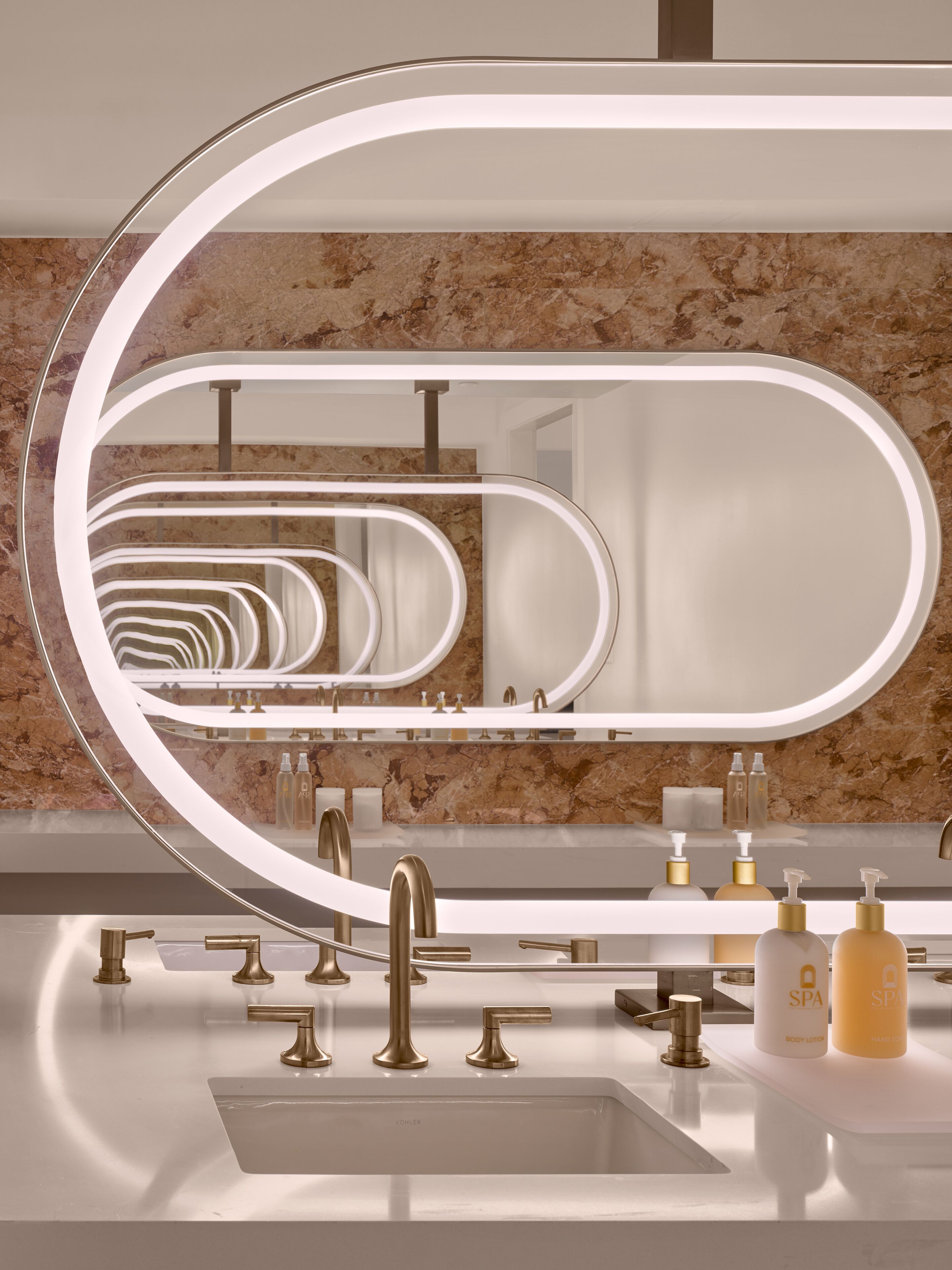 A modern bathroom with a large oval mirror, gold faucets, and a marble backsplash.