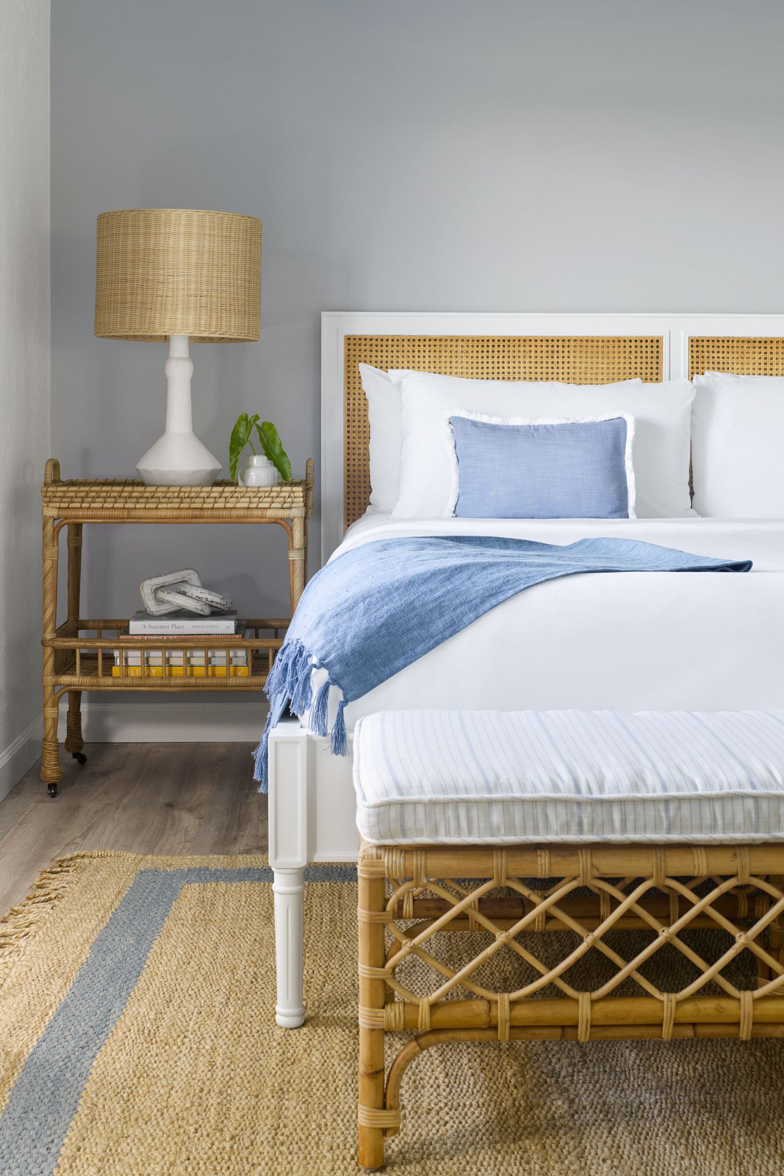 A neatly arranged bedroom with a bed, bedside table, lamp, and a small shelf with books and a plant.