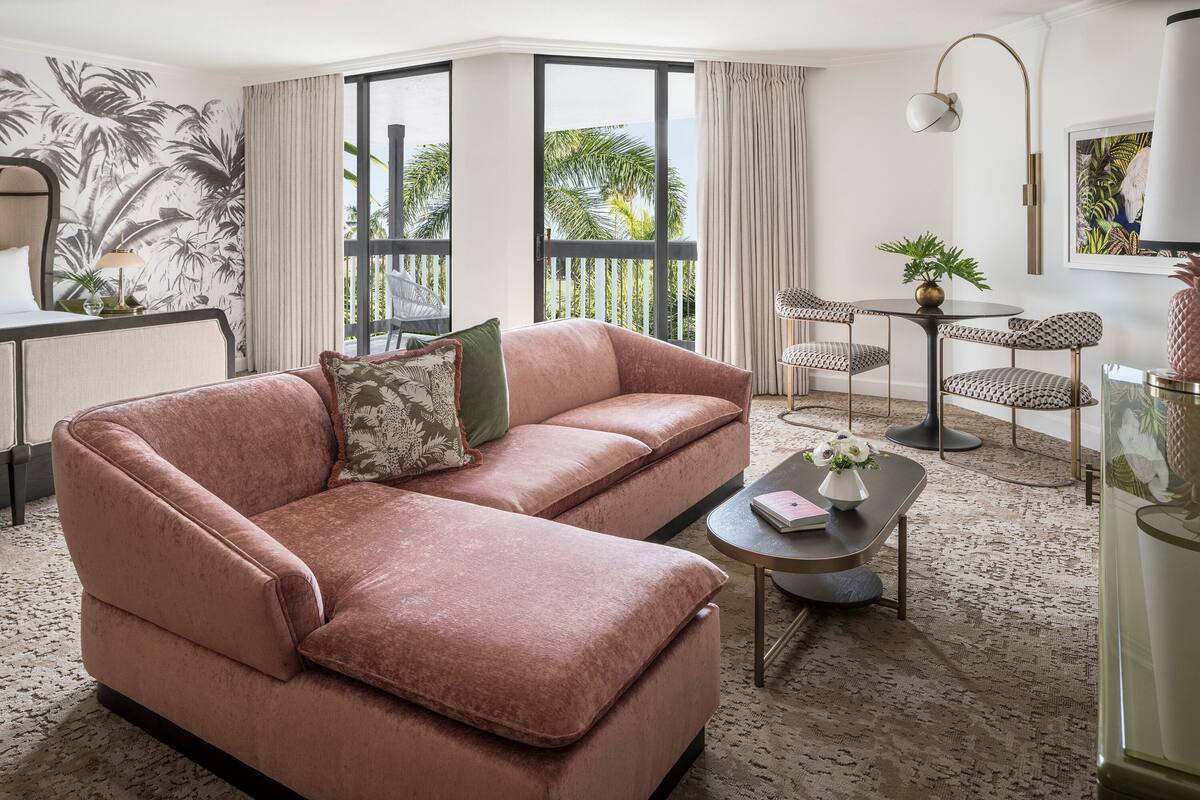 Een moderne woonkamer met een roze bank, een kleine ronde tafel met een bloevas, en een groot raam met uitzicht op palmbomen.