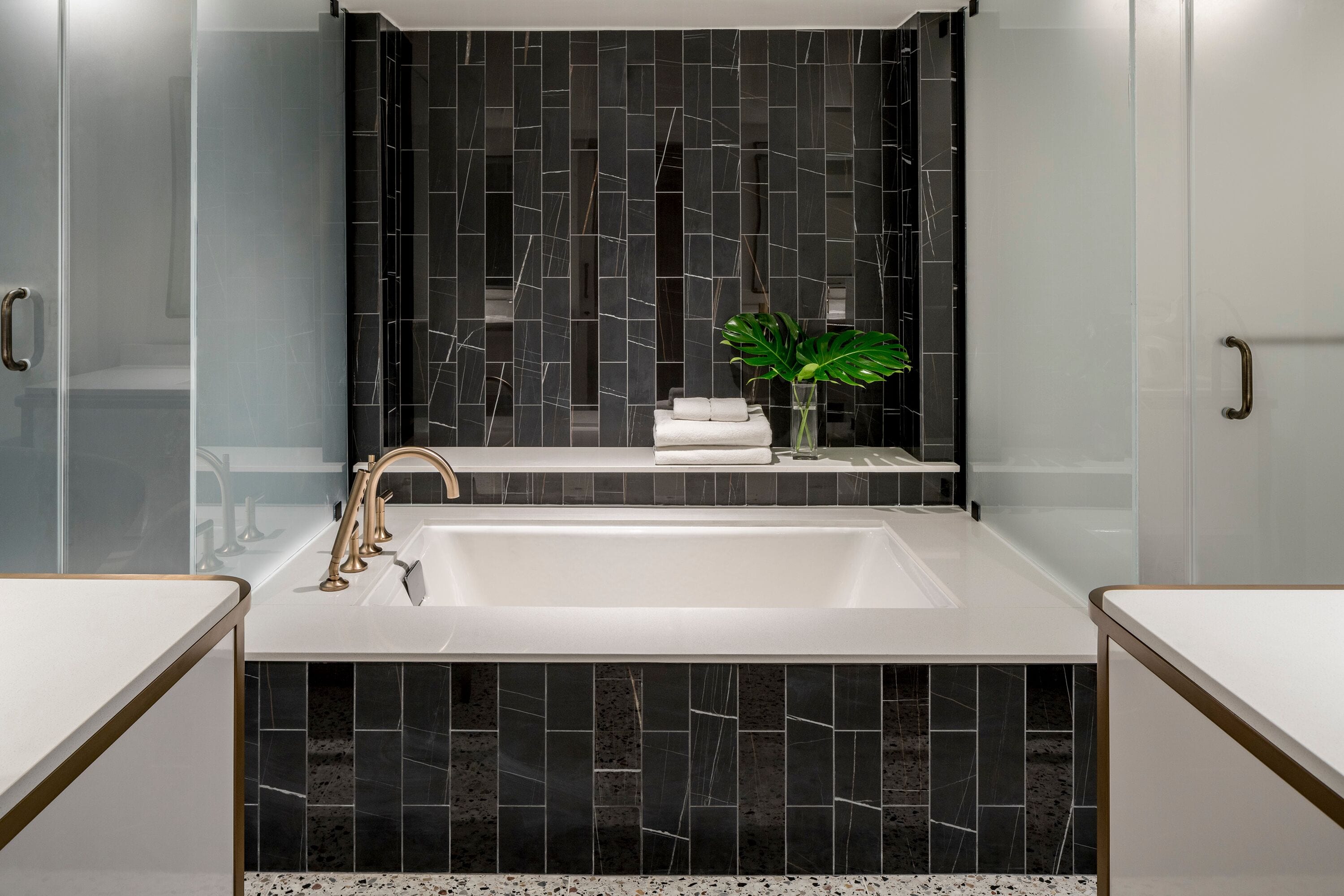 Un baño moderno con una bañera blanca grande, azulejos de mármol negro y un estante pequeño con toallas y una planta.