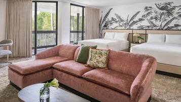 A hotel room with a pink sectional sofa, a bed, a nightstand with a lamp, and a large tropical mural on the wall.