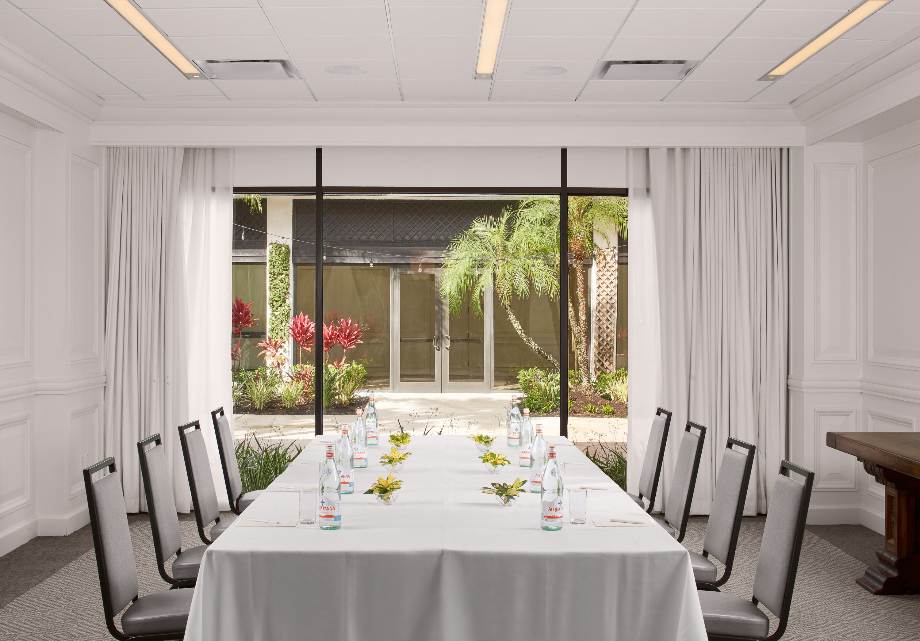A conference room with a long table, chairs, water bottles, and a view of a garden through large windows.