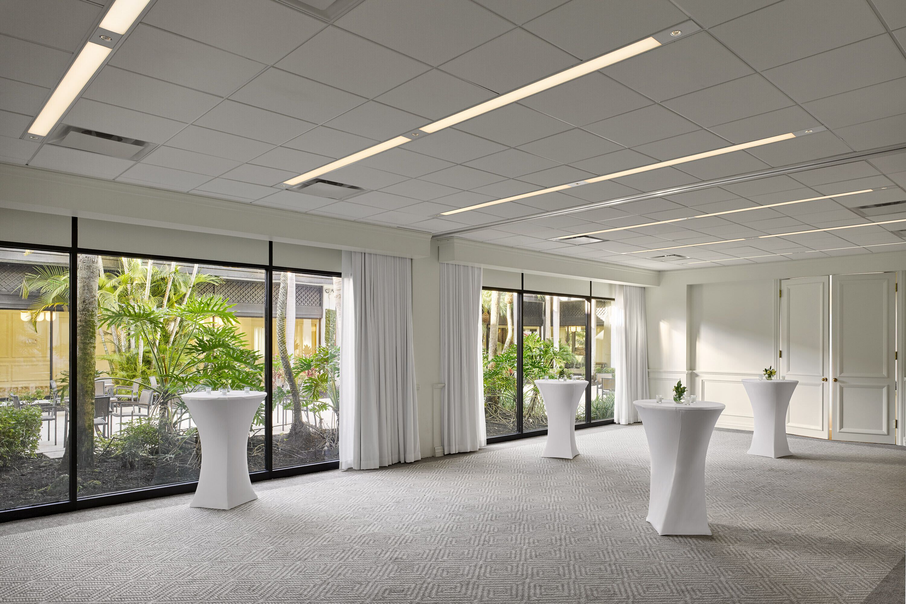 A spacious room with large windows, white curtains, and multiple white round tables.