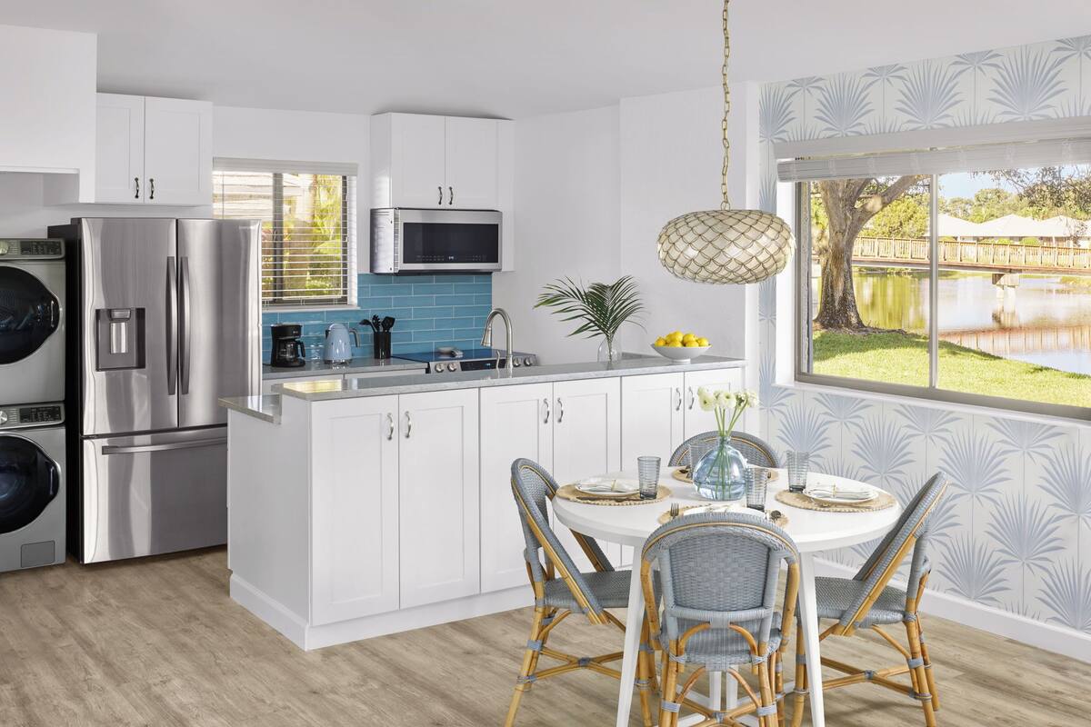 Een moderne keuken met witte kastjes, roestvrijstalen apparatuur en een eethoek met rieten stoelen en een ronde tafel.
