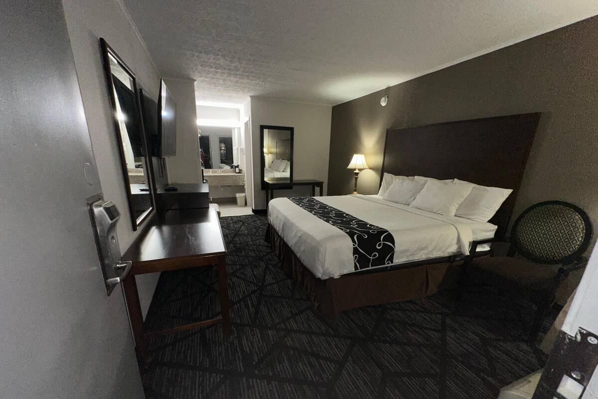 A hotel room with a bed, a desk, a chair, a TV, a mirror, and a bathroom visible in the background.