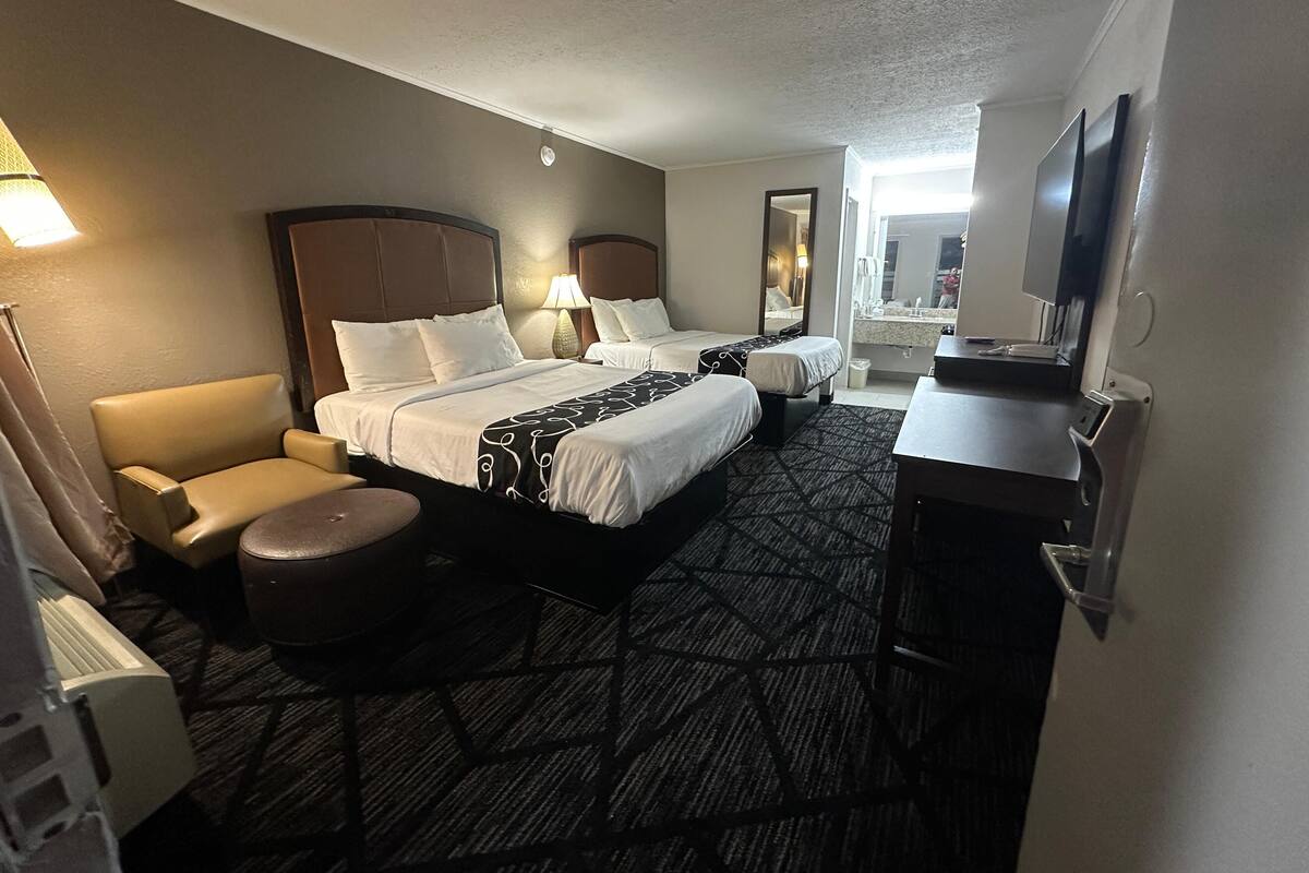 A hotel room with two beds, a desk, a chair, a TV, and a bathroom visible through an open door.