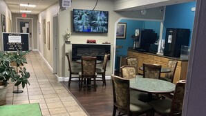 A hotel lobby with a reception desk, a television mounted on the wall, a dining area with wooden chairs and a green table, and a tiled floor.