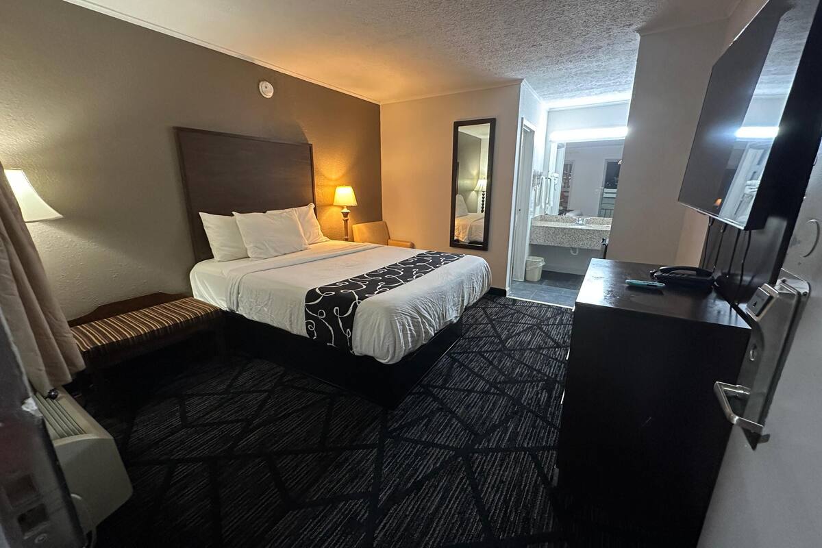 A hotel room with a bed, bedside lamp, dresser, and a mirror.