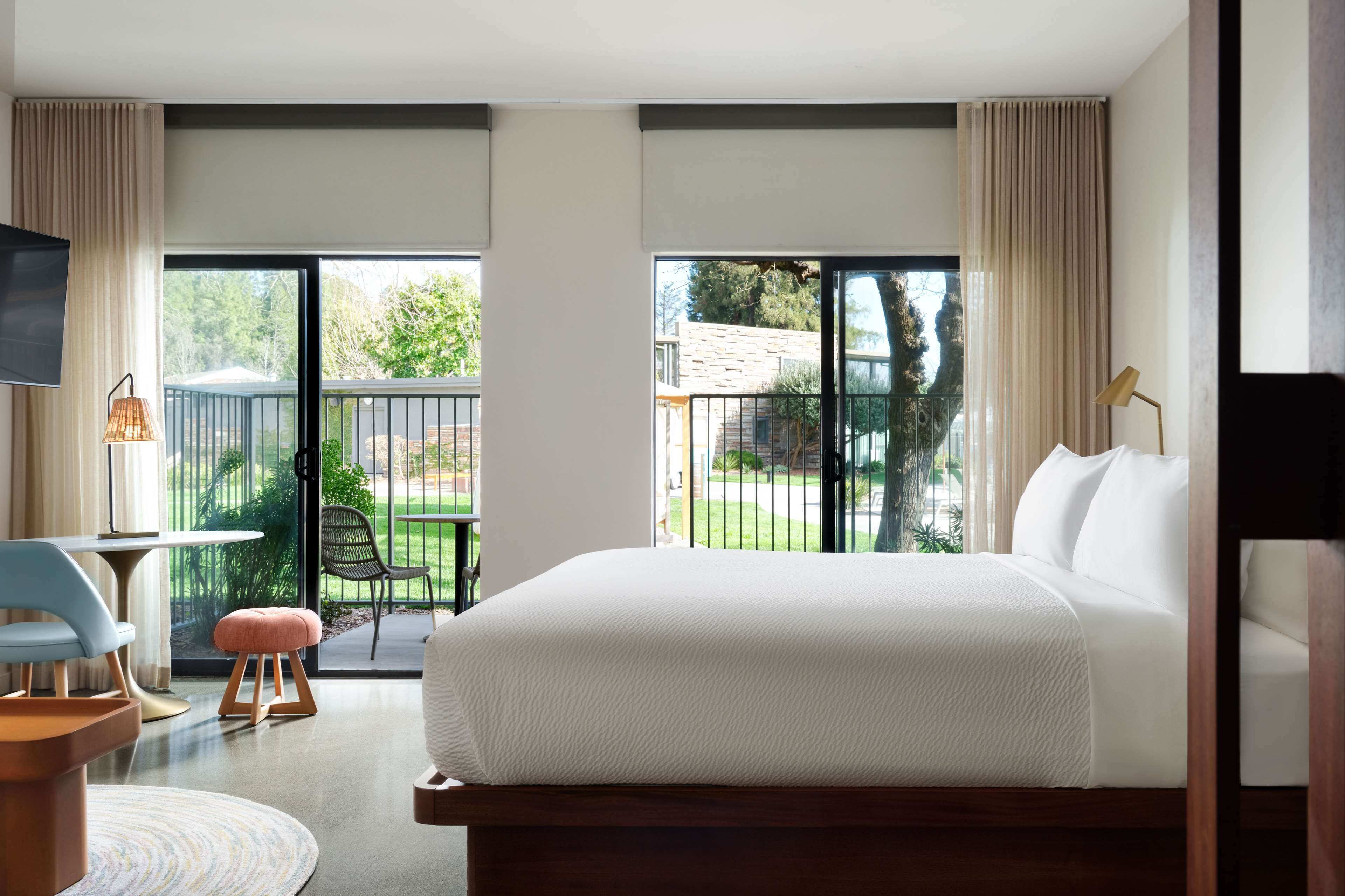 A hotel room with a large bed, a TV, a desk with a chair, and a balcony with a view of the outdoors.