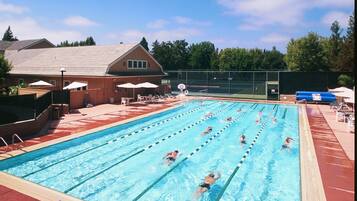 A swimming pool with multiple lanes, surrounded by a fence, with a building and trees in the background.