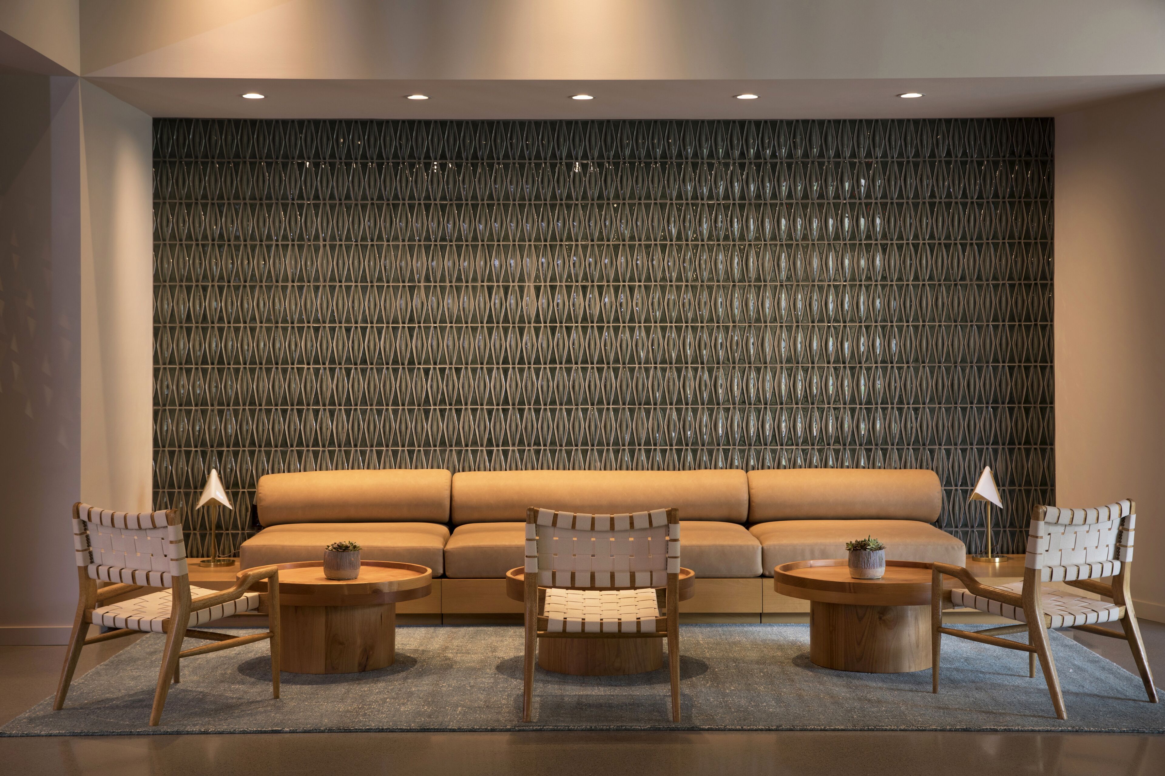 A modern living room with a patterned wall, a beige sofa, two wooden chairs, and two round wooden tables.
